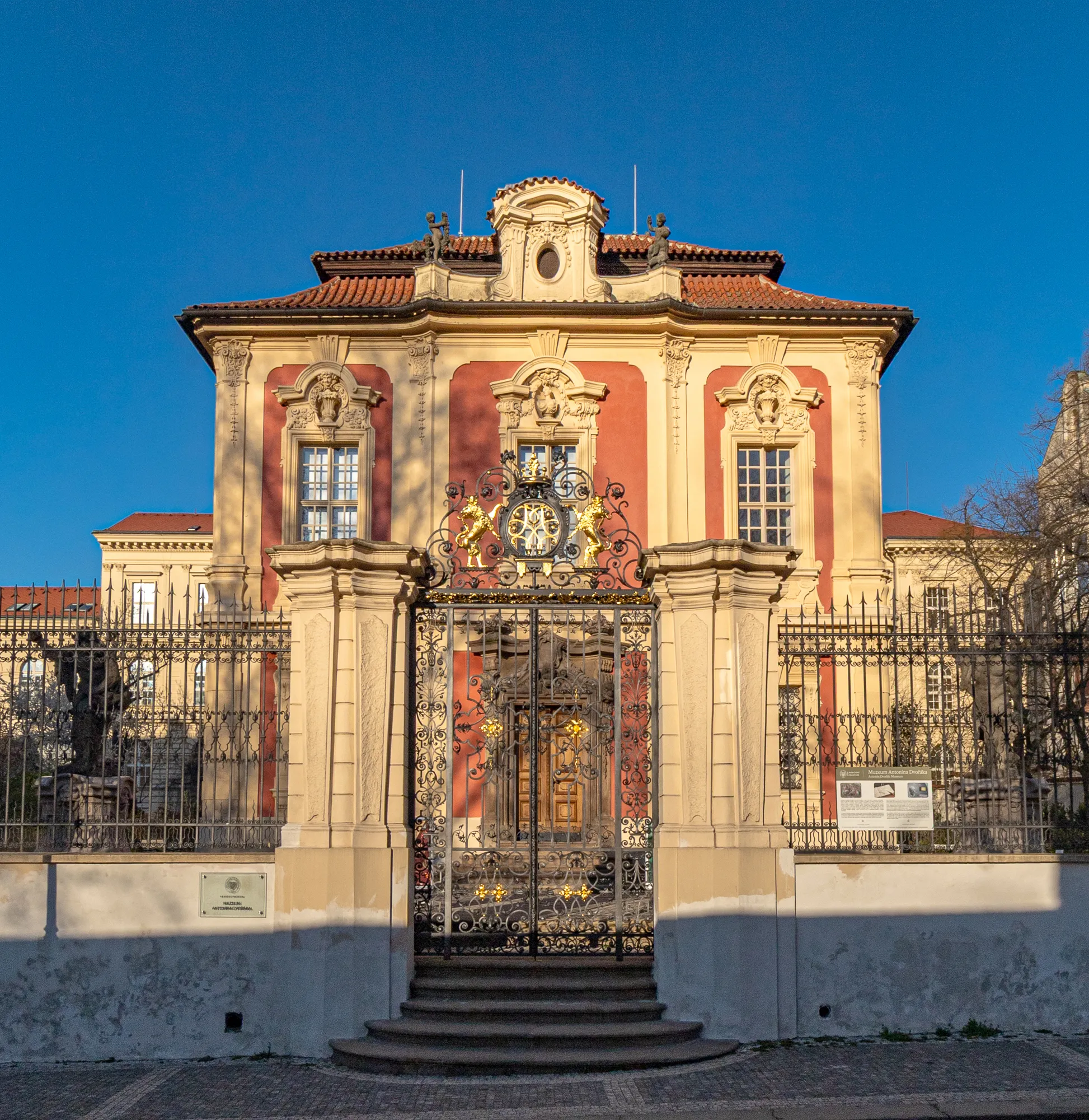 Budynek w stylu barokowym z czerwoną i beżową fasadą, ozdobną bramą i schodami prowadzącymi do wejścia pod błękitnym niebem