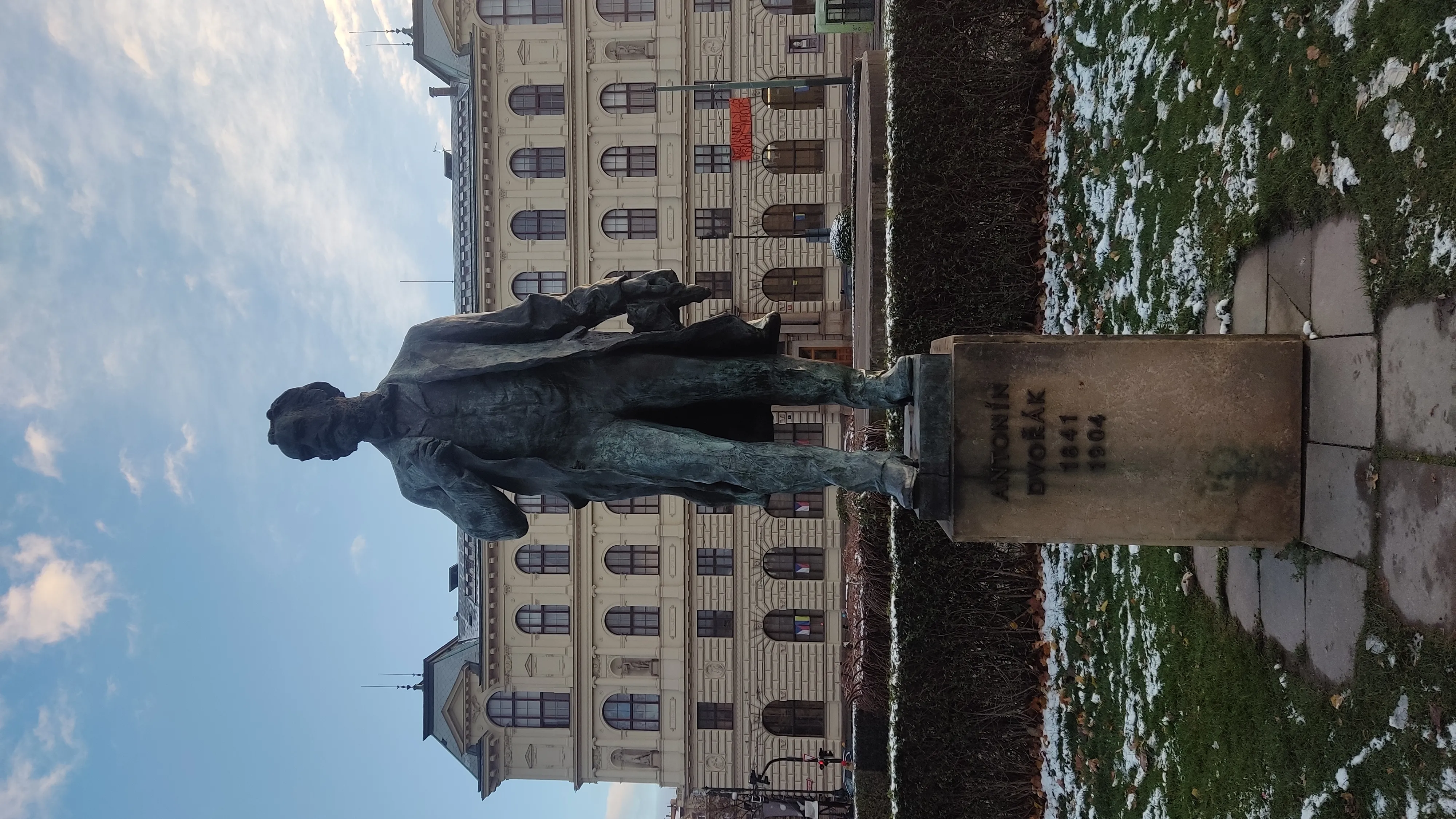 Brązowy posąg Antonína Dvořáka przed dużym budynkiem ze śniegiem na ziemi
