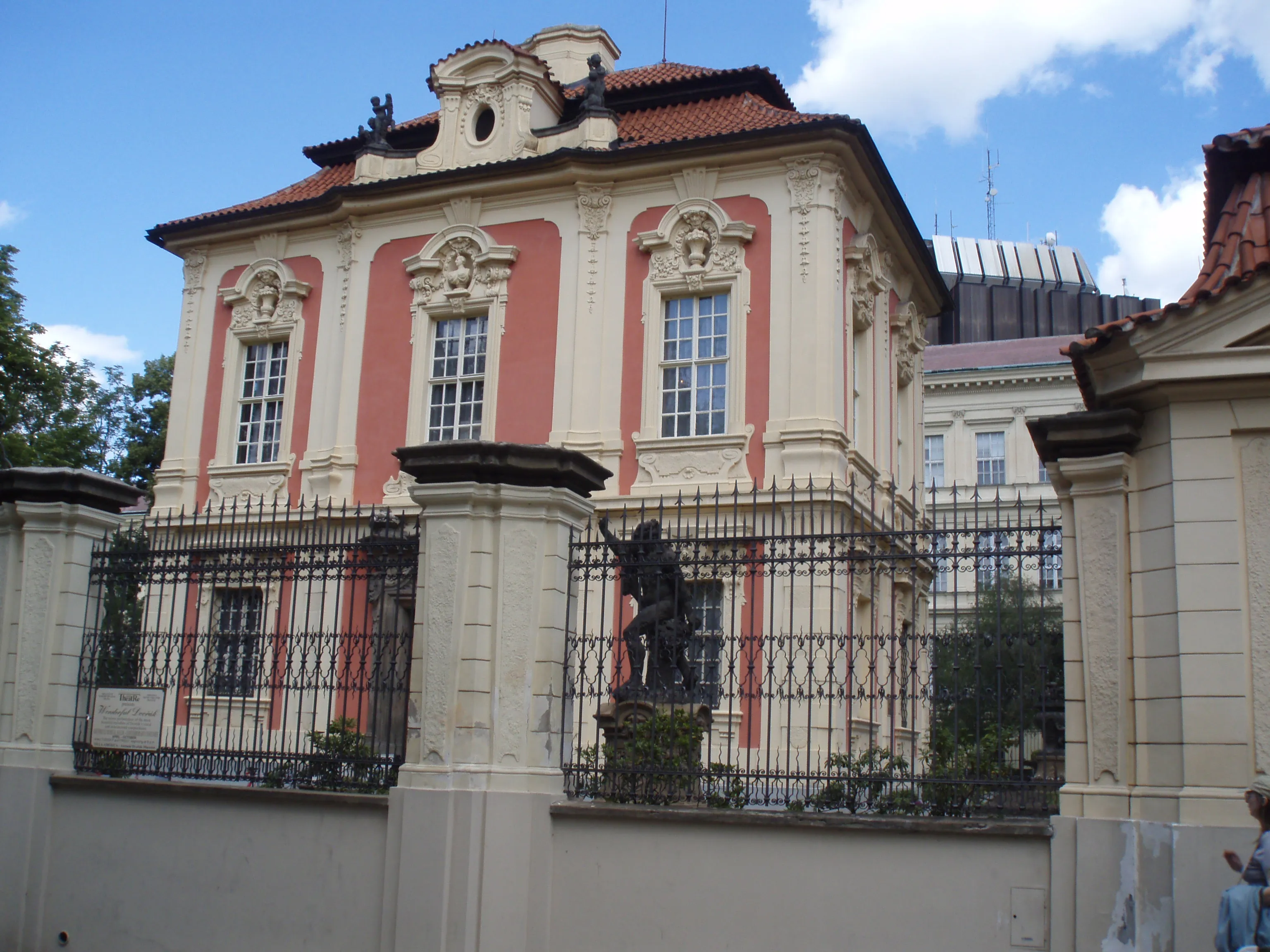 Budynek w stylu barokowym z różowo-kremową fasadą, dachem z czerwonej dachówki i czarnym żelaznym ogrodzeniem.