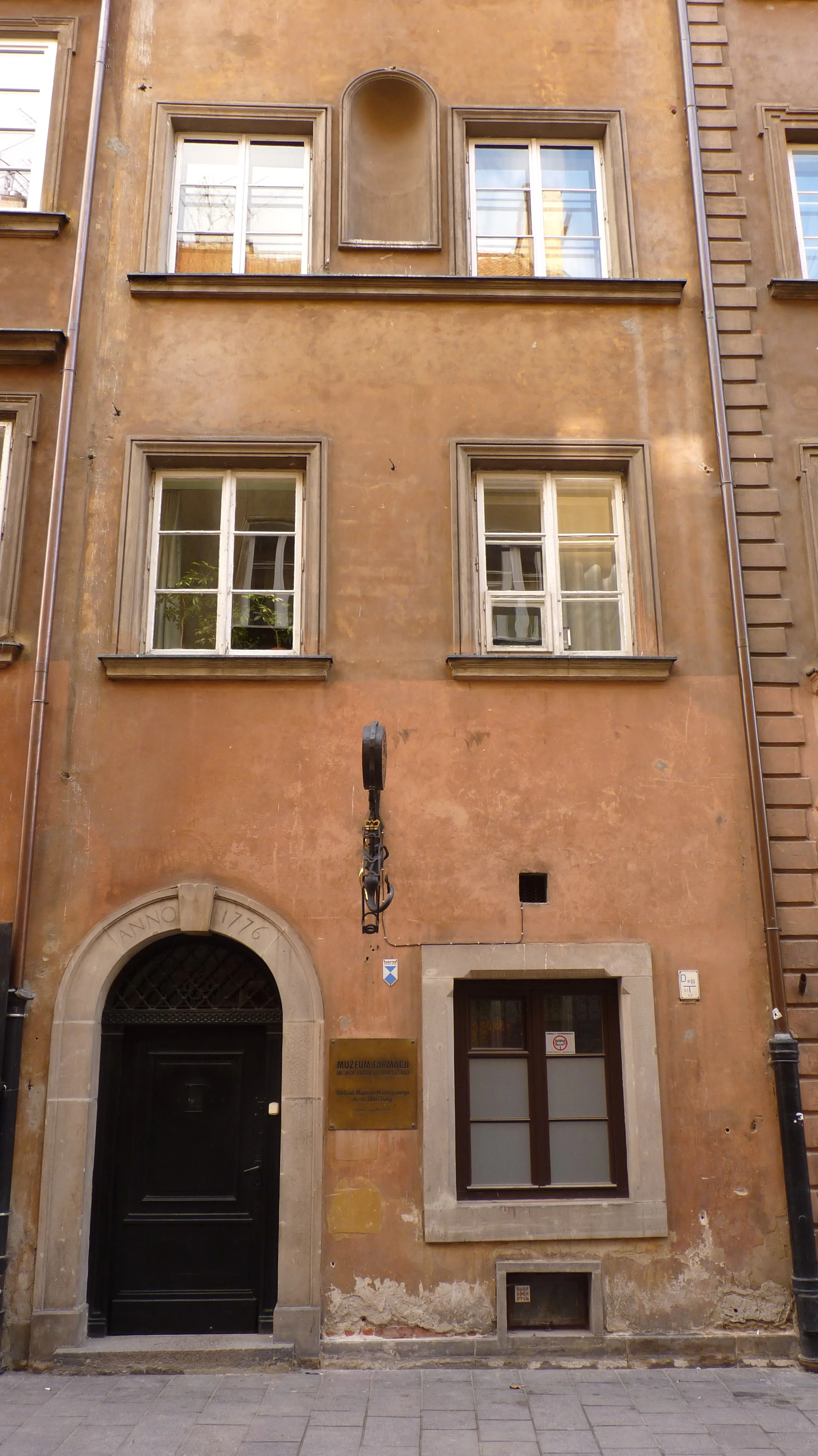 Een historisch gebouw met een terracotta gevel, zwarte boogdeur en meerdere ramen, met een plaquette en apothekersbord naast de ingang.