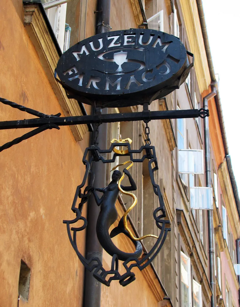 Zwart smeedijzeren uithangbord met de tekst 'MUZEUM FARMACJI' en een medisch symbool, hangend aan een gebouwmuur.