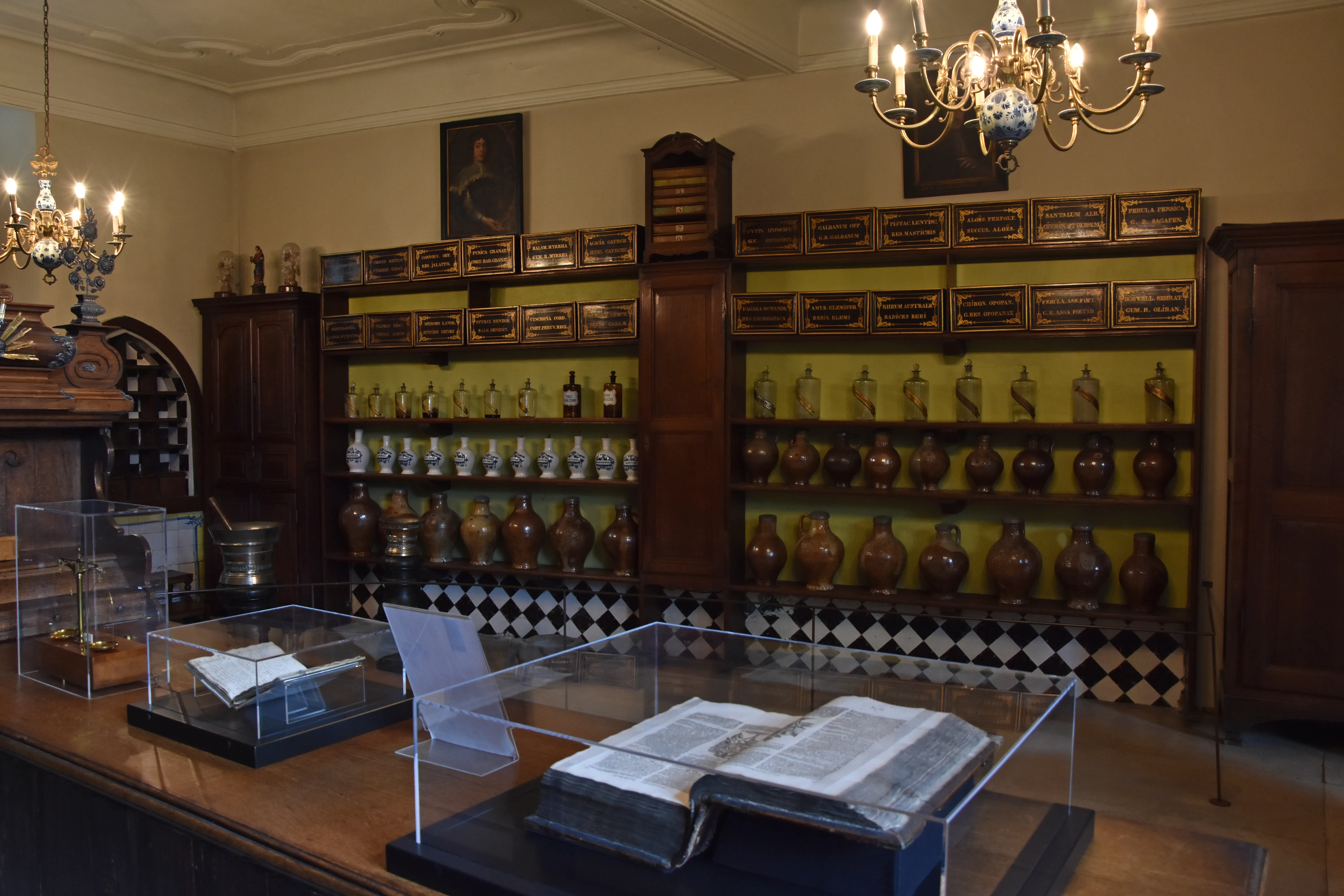 Interior de una farmacia histórica con estantes de madera altos llenos de hileras de frascos de cerámica marrón, lámparas de araña ornamentadas en el techo y vitrinas de cristal con libros antiguos en primer plano.