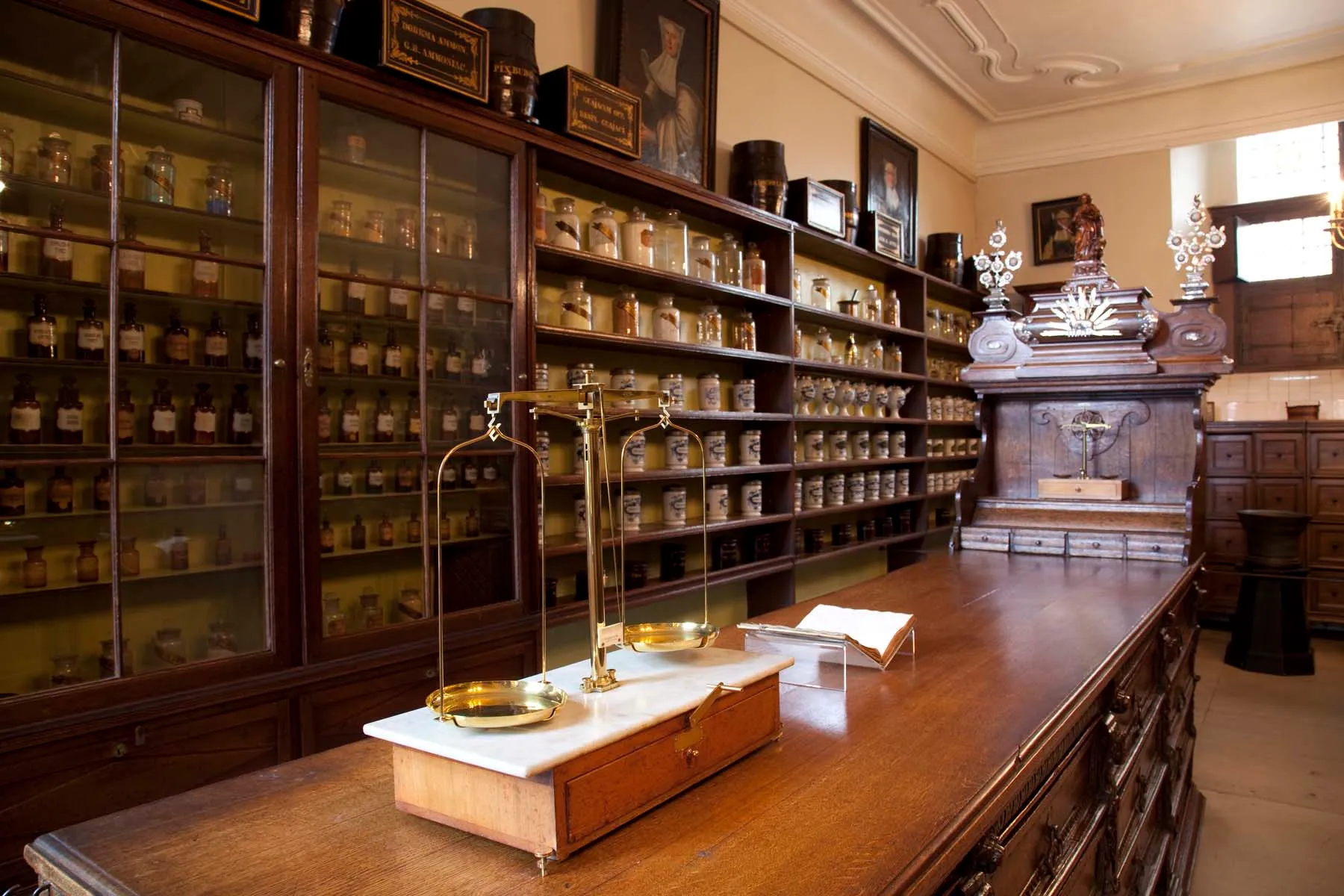 Interior de una farmacia histórica con altos armarios de madera llenos de hileras de frascos de cerámica medicinal, un largo mostrador de madera con una balanza de latón sobre una encimera de mármol y muebles de madera tallada ornamentada al fondo.