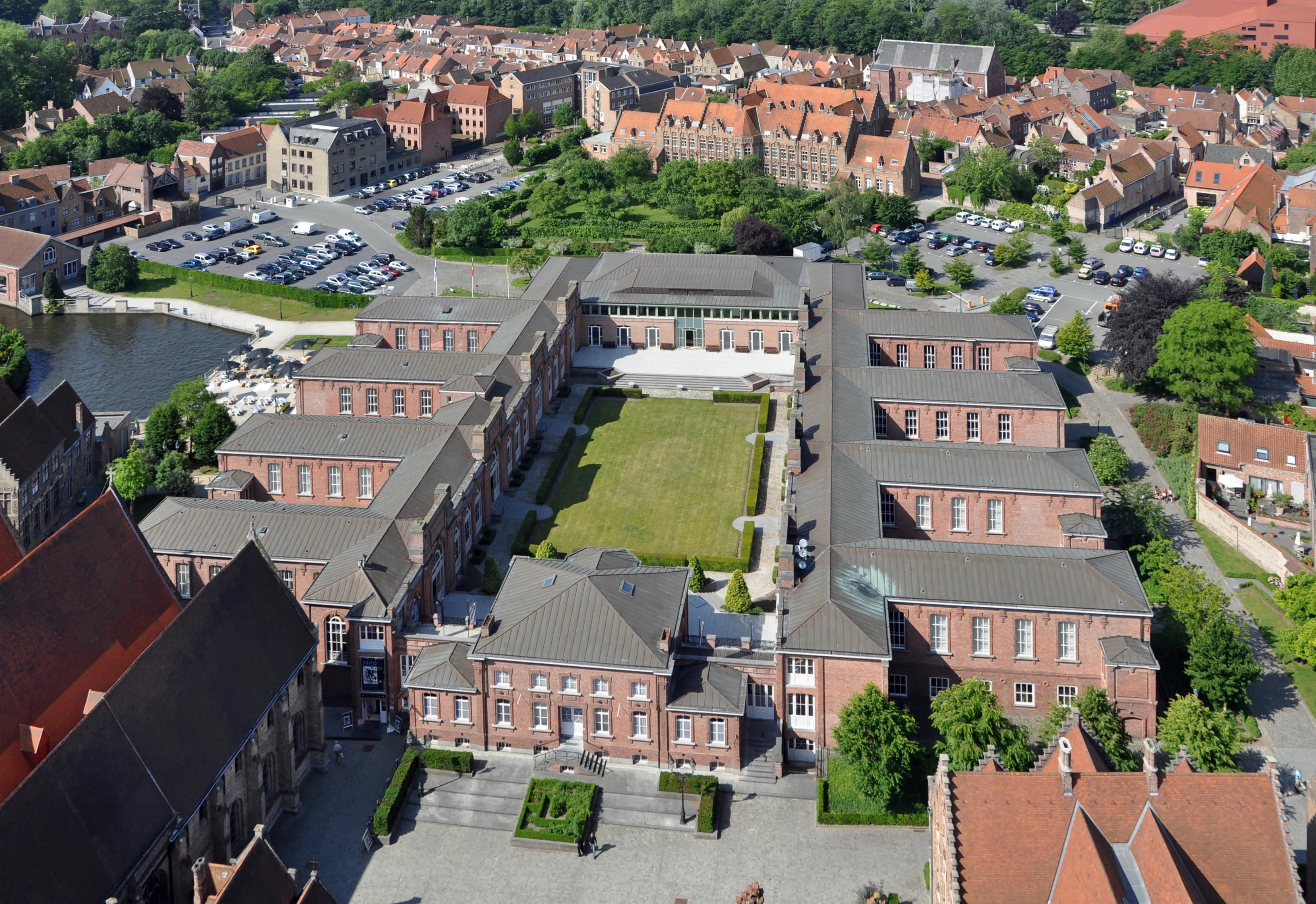 Vista aérea del complejo de ladrillo del Sint-Janshospitaal con un patio verde, rodeado por el paisaje urbano de Brujas con su canal, árboles y zonas de aparcamiento.