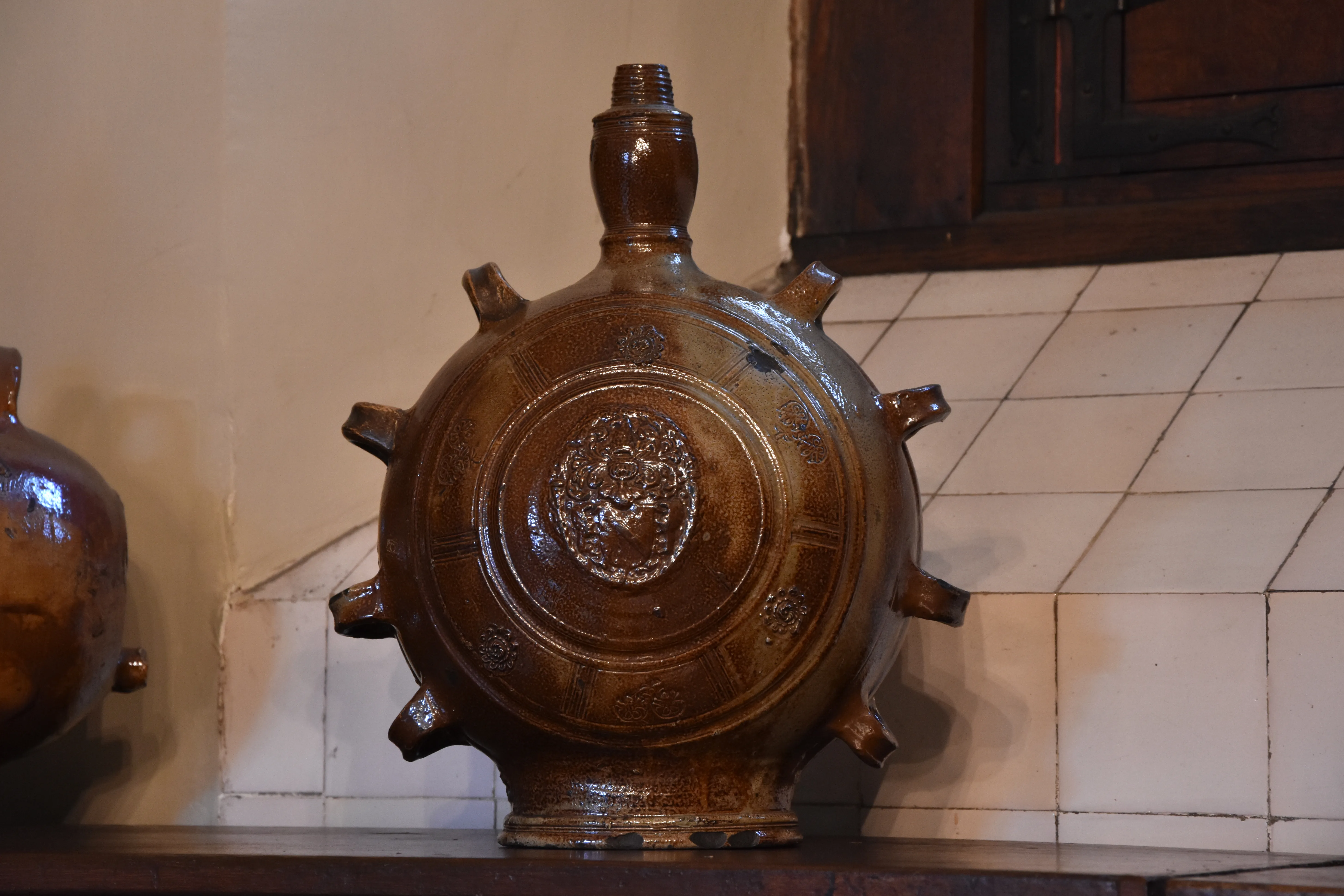 A large round brown stoneware medicinal jar with multiple small protruding spouts around its circumference and a narrow neck, displayed on a wooden shelf against a white tiled wall.