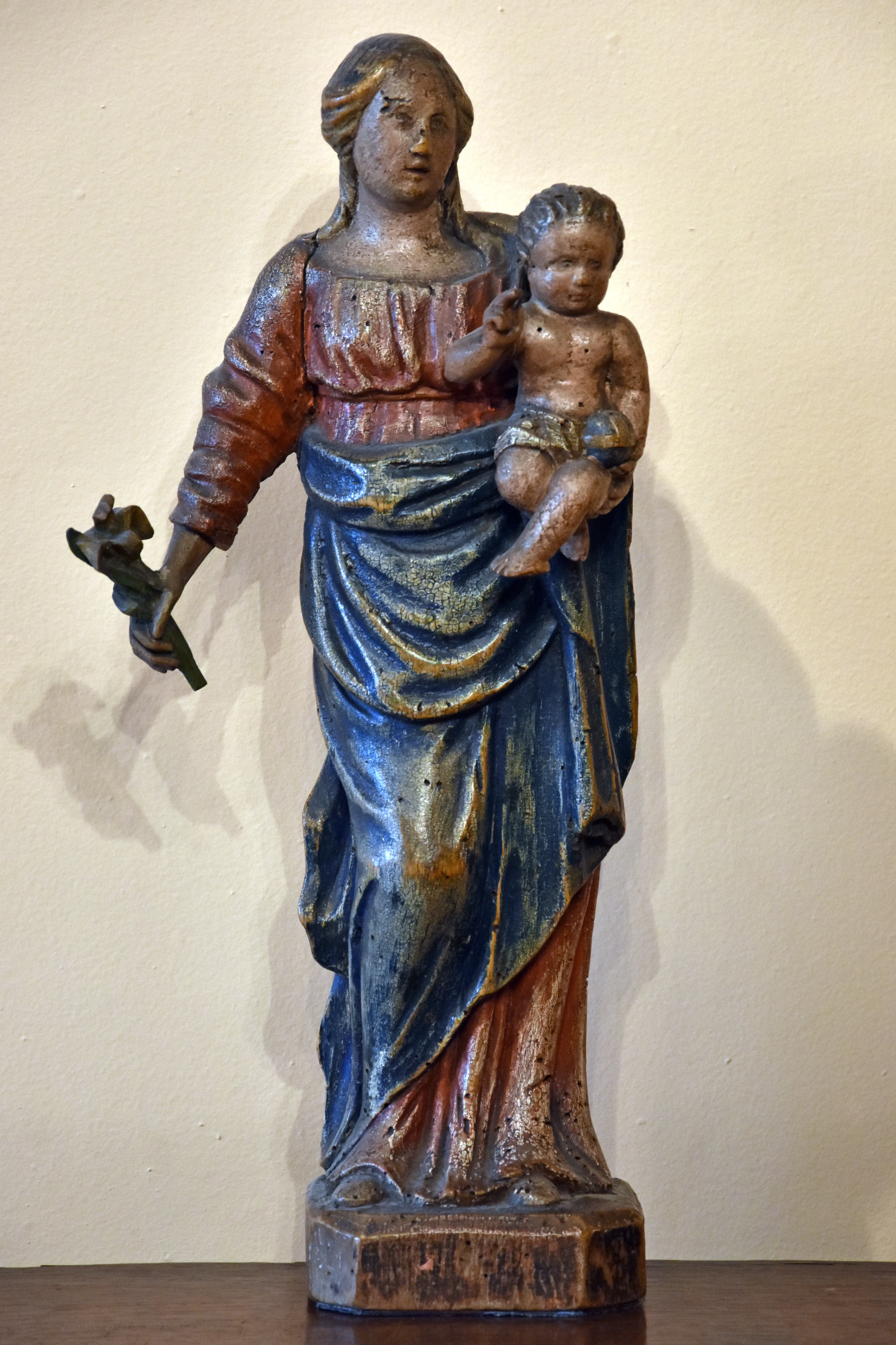 A polychrome wooden statue of the Virgin Mary holding the Christ child in her left arm and a flower in her right hand, standing on a hexagonal base against a plain wall.