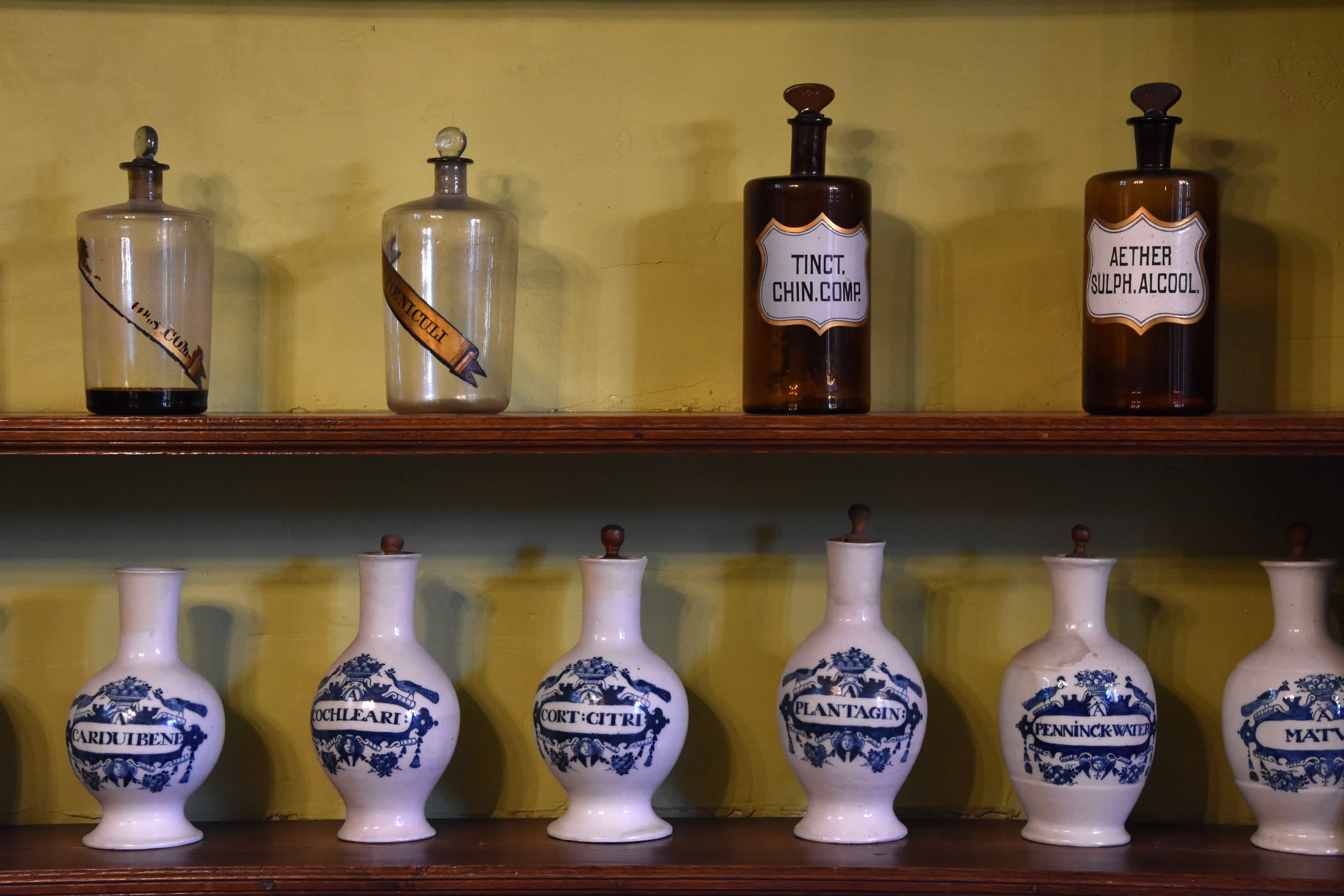 Dos estantes de madera que sostienen botellas de vidrio antiguas y frascos de farmacia de cerámica azul y blanca con etiquetas escritas a mano.