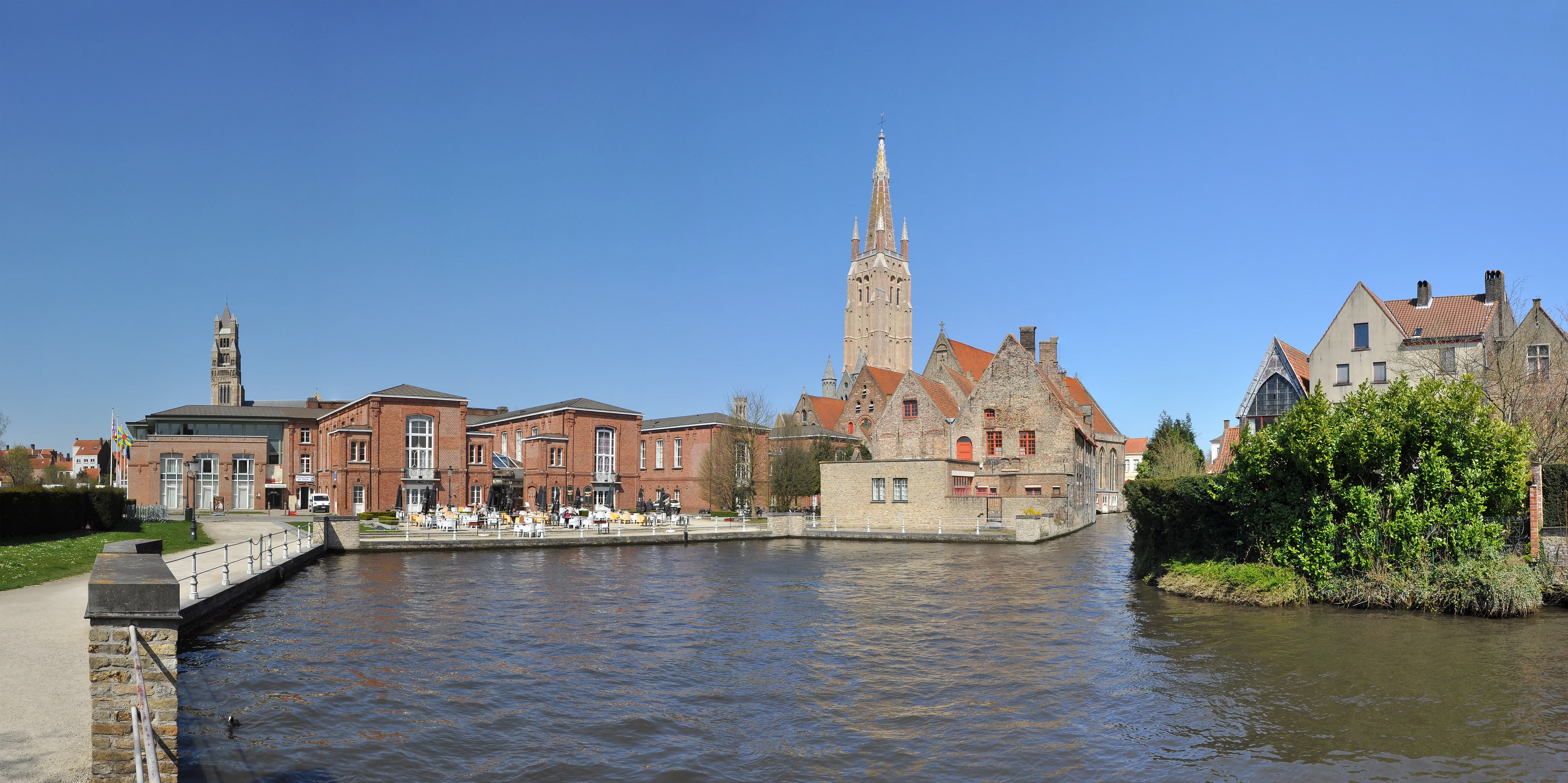 Vista panorámica del complejo del Sint-Janshospitaal a lo largo de un canal en Brujas, con edificios históricos de ladrillo y la torre de una iglesia bajo un cielo azul despejado.