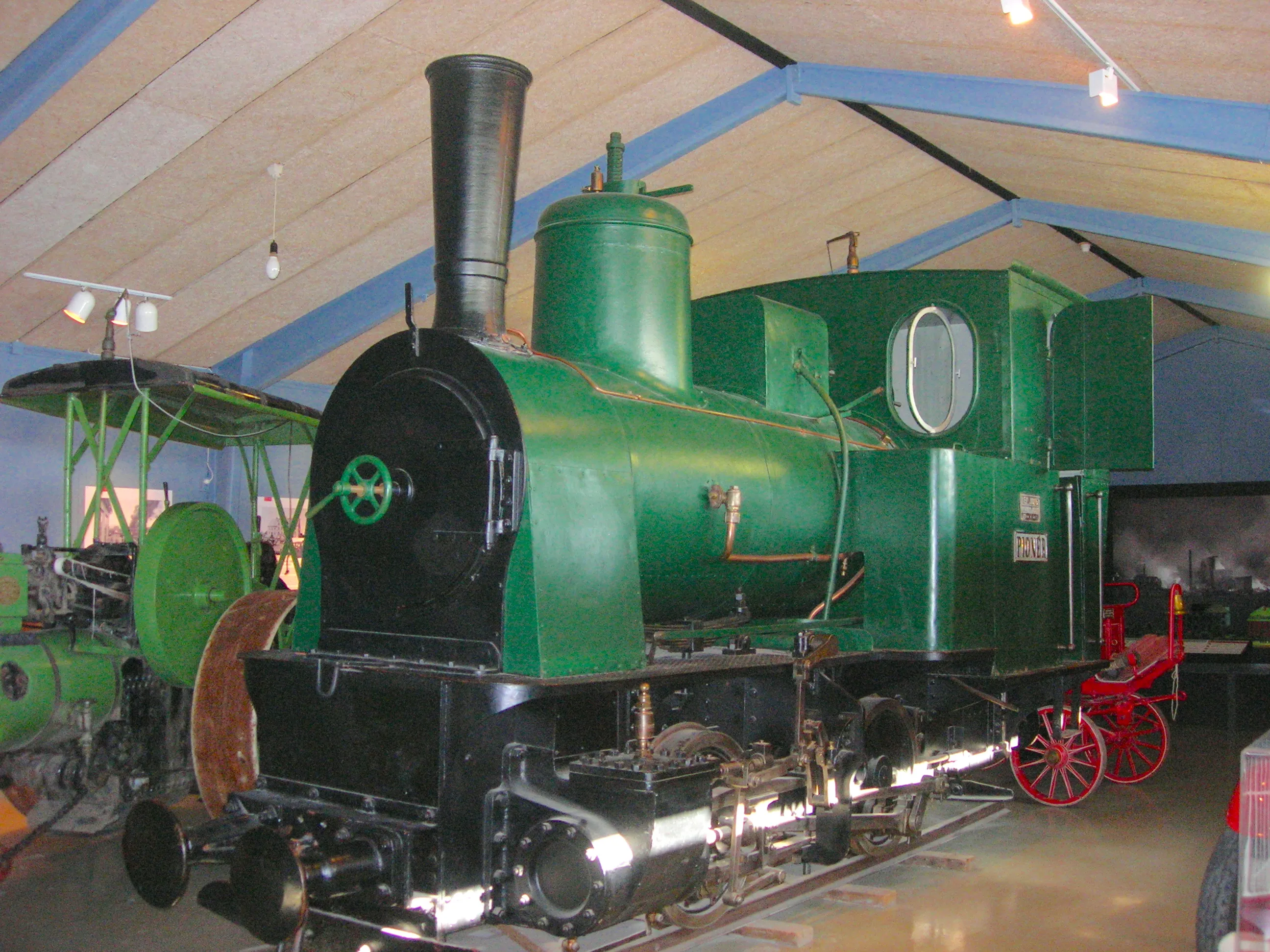 A green steam locomotive with a tall black smokestack on display inside a museum hall, with a small red-wheeled cart beside it and blue ceiling beams overhead.