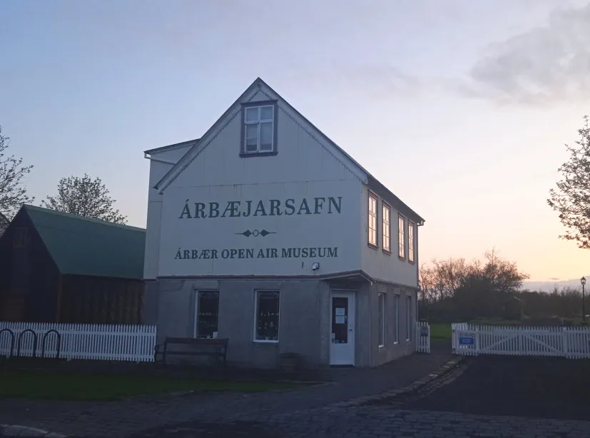 White two-story gabled building with ÁRBÆJARSafn and ÁRBÆR OPEN AIR MUSEUM signage on the facade, surrounded by a white picket fence at dusk