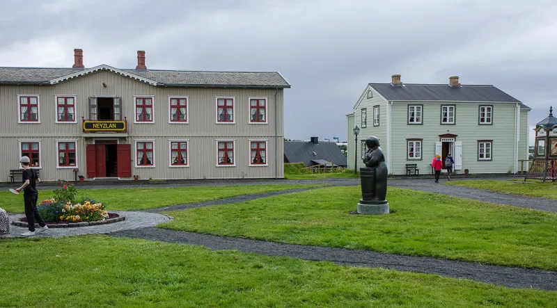 Two historic buildings with red-trimmed windows and a grassy courtyard featuring a bronze bust statue.