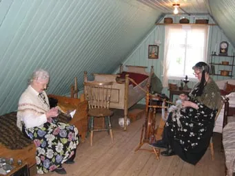 Two women in historic clothing inside a rustic attic room with a slanted ceiling, one operating a spinning wheel while the other sits on a wooden bed.