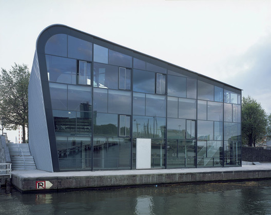 Nowoczesny szklany budynek z zakrzywionym dachem nad wodą, odbijający niebo i otoczenie. Budynek ARCAM (Amsterdam Centre for Architecture).