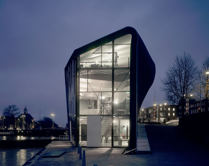 Nowoczesny budynek ze szkła i stali z zakrzywioną fasadą o zmierzchu, odbijający się w wodzie. Amsterdamskie Centrum Architektury (ARCAM) oświetlone w nocy.
