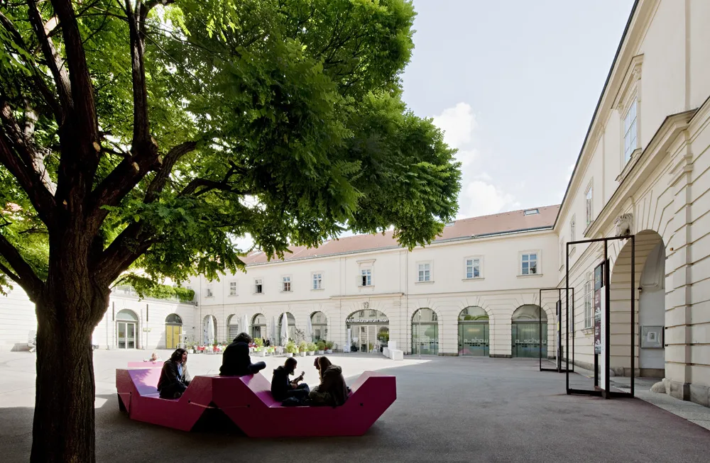 Courtyard with a large tree, a modern pink bench with people, and the white facade of the Architekturzentrum Wien building.