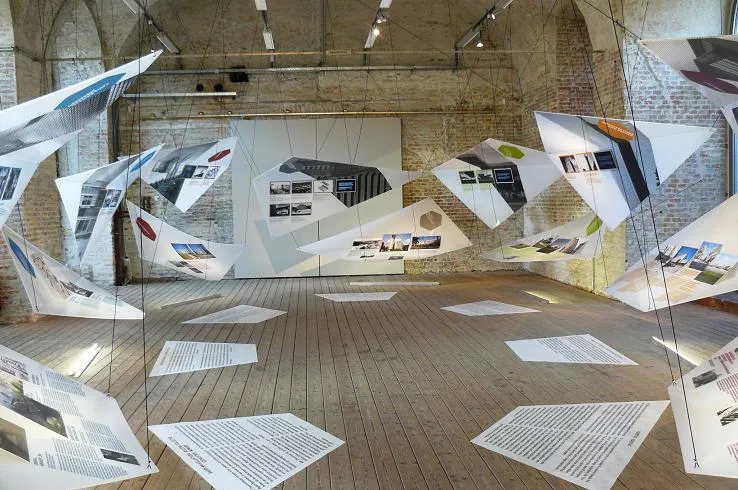 Suspended architectural drawings and floor-placed documents in an exhibition space with brick walls and wooden floors