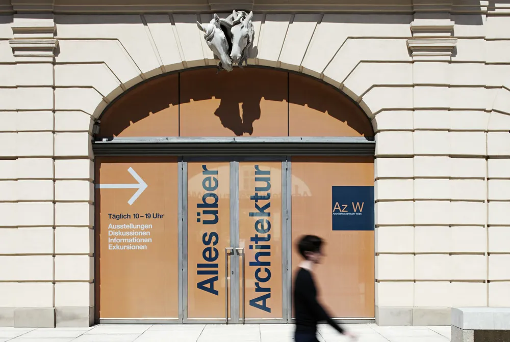Arched entrance to a building with glass doors displaying 'Alles über Architektur' and 'Az W' signs, a person walking in front