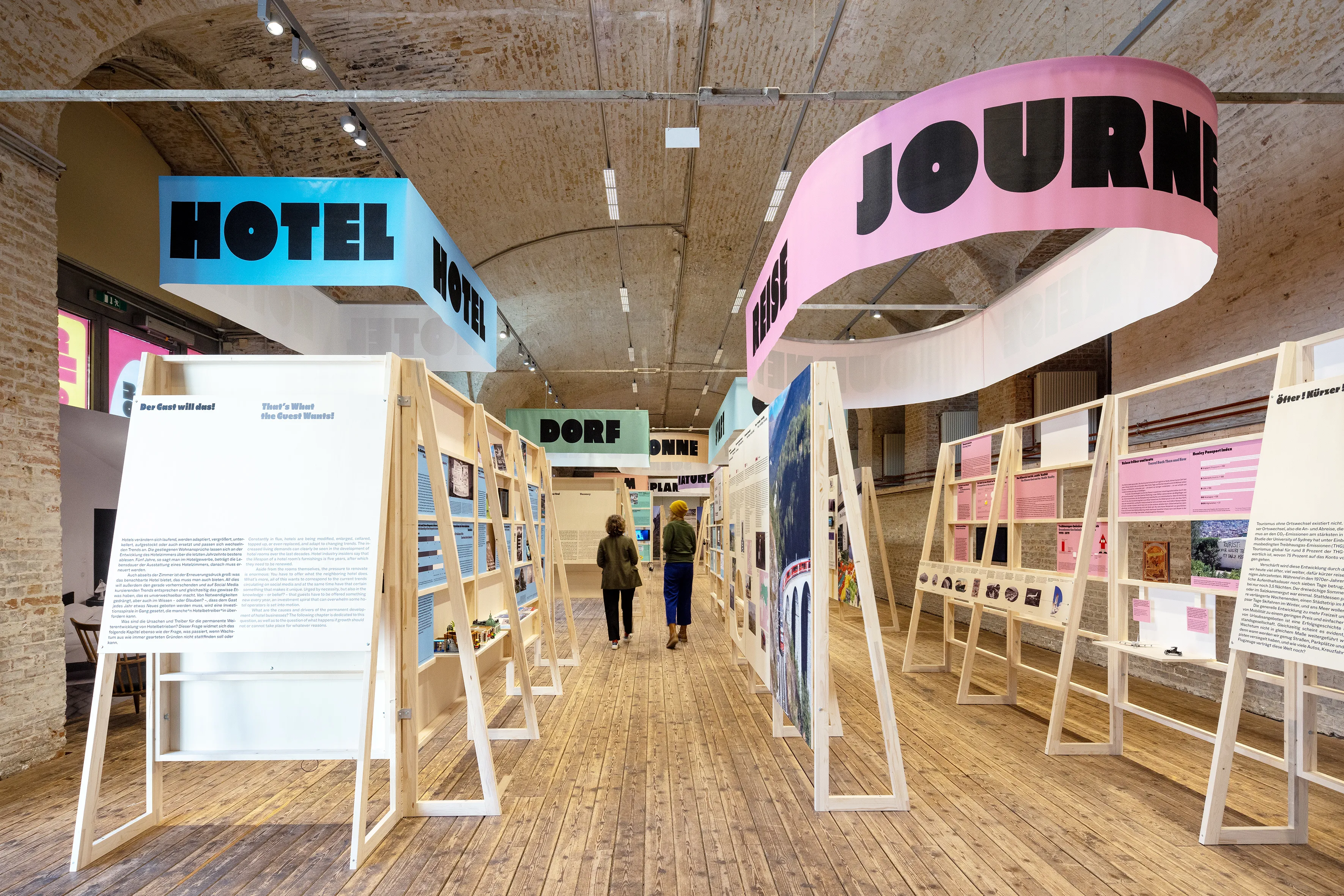 Exhibition space with large text installations reading HOTEL and JOURNEY, display boards, and wooden structures on a wooden floor.