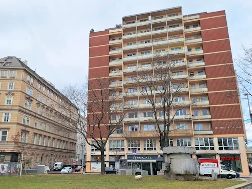 A tall brick building with balconies and large windows, surrounded by trees and other urban structures.