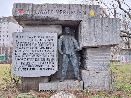 Stone memorial with a statue of a man and an inscription plaque, set outdoors with buildings in the background