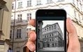 A hand holding a smartphone showing a black and white photo of a building, with the same building visible in the background