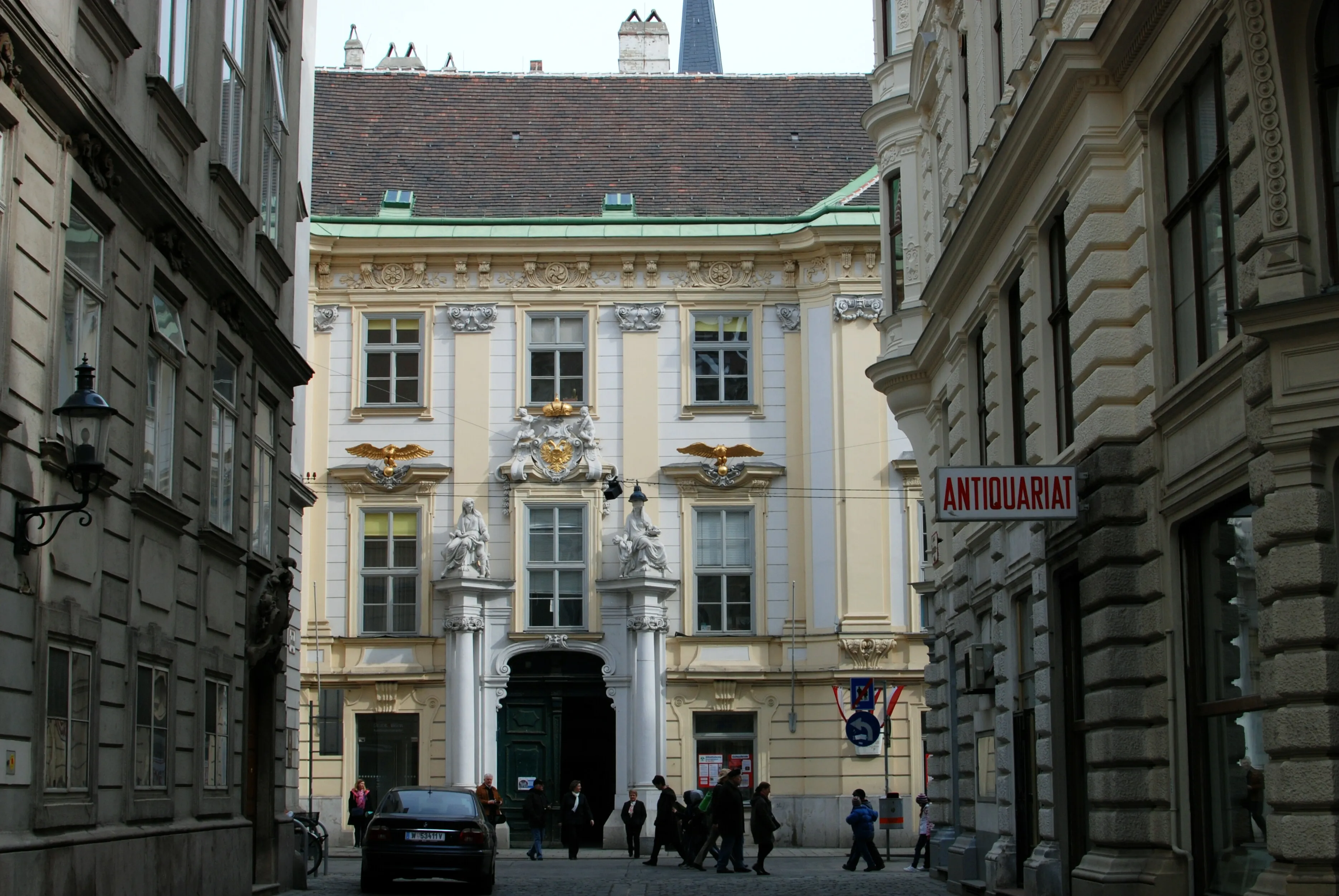 Historic building with classical architecture on a narrow street, featuring a central entrance with columns and ornate decorations, people walking in front, and a sign reading 'ANTIQUARIAT' on the right side