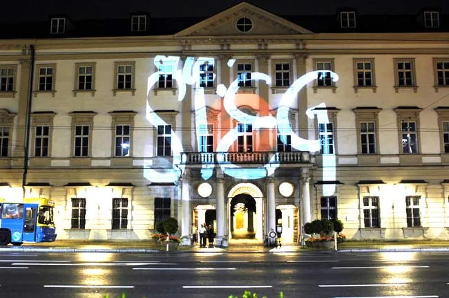 Vista nocturna de la fachada del edificio ARGEkultur Salzburg con una gran proyección de luz blanca que dice 'ARGE' sobre la arquitectura clásica, con un autobús azul visible en la calle.