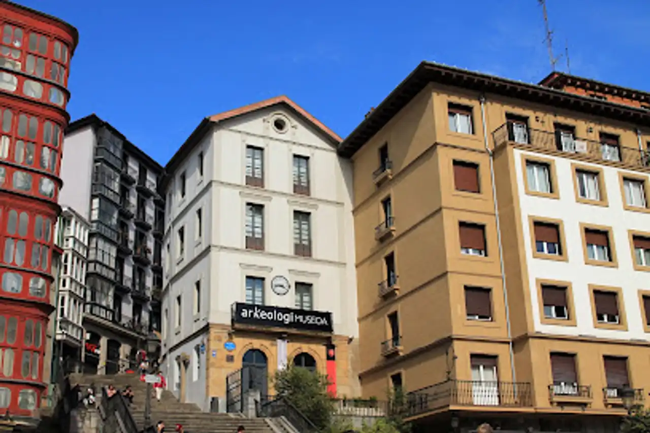 Edificio blanco con el letrero 'arkeologiamuseoa' flanqueado por edificios rojos y beige bajo un cielo azul despejado, con personas en la escalinata frontal