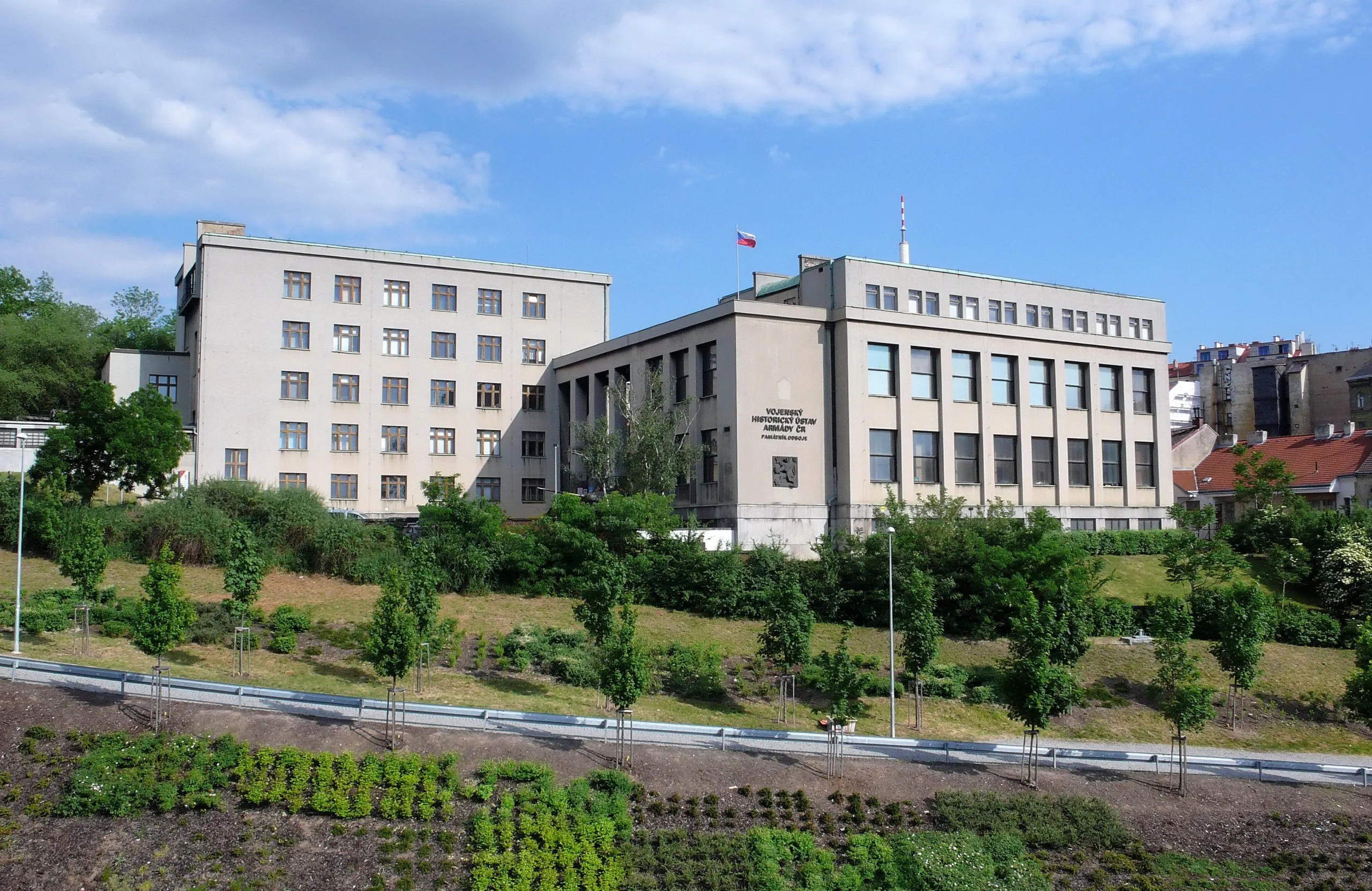 Een groot beige gebouw met veel ramen, geïdentificeerd als het Legermuseum Žižkov, gelegen op een grasrijke heuvel met bomen en een blauwe lucht op de achtergrond.