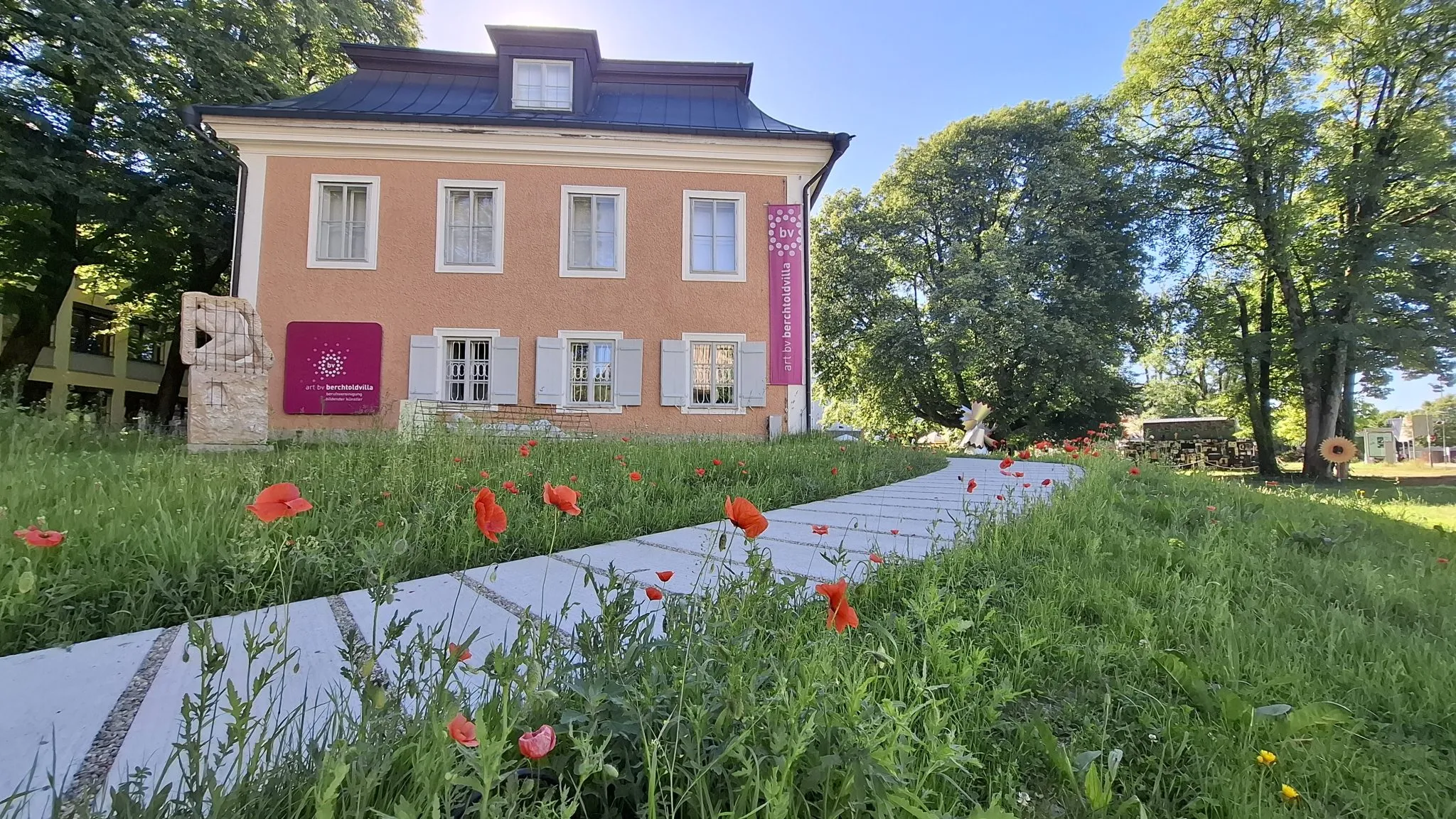 Edificio de villa histórica rosa con un sendero de piedra blanca y amapolas rojas en el jardín delantero, con pancartas de exposición en color magenta en la fachada