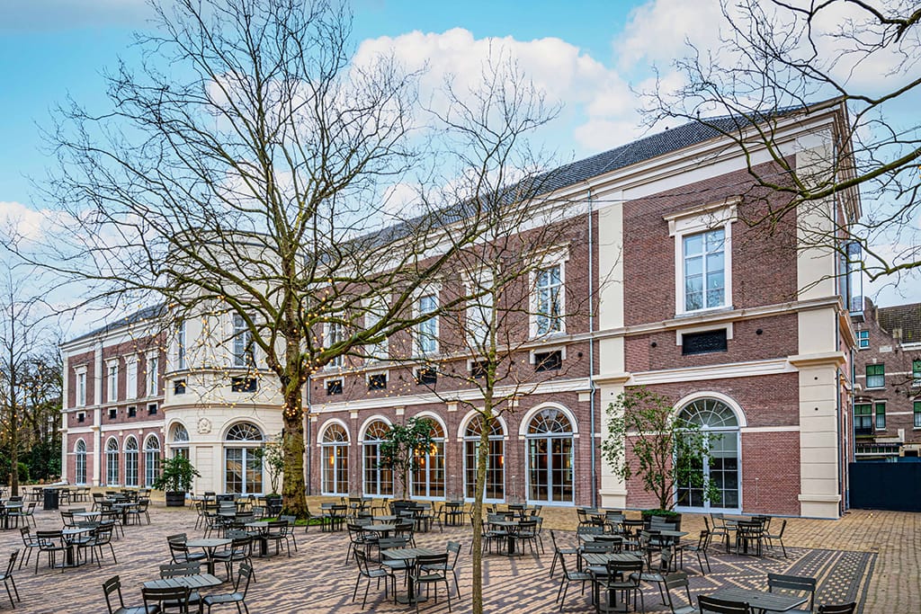 Brick building with arched windows and outdoor seating area in courtyard. Trees without leaves under blue sky with clouds.
