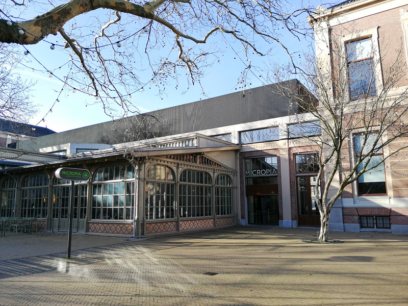 Buitenkant van het ARTIS Micropia museumgebouw met 'MICROPIA' uithangbord boven de ingang, glazen pui en kale bomen op een zonnige dag