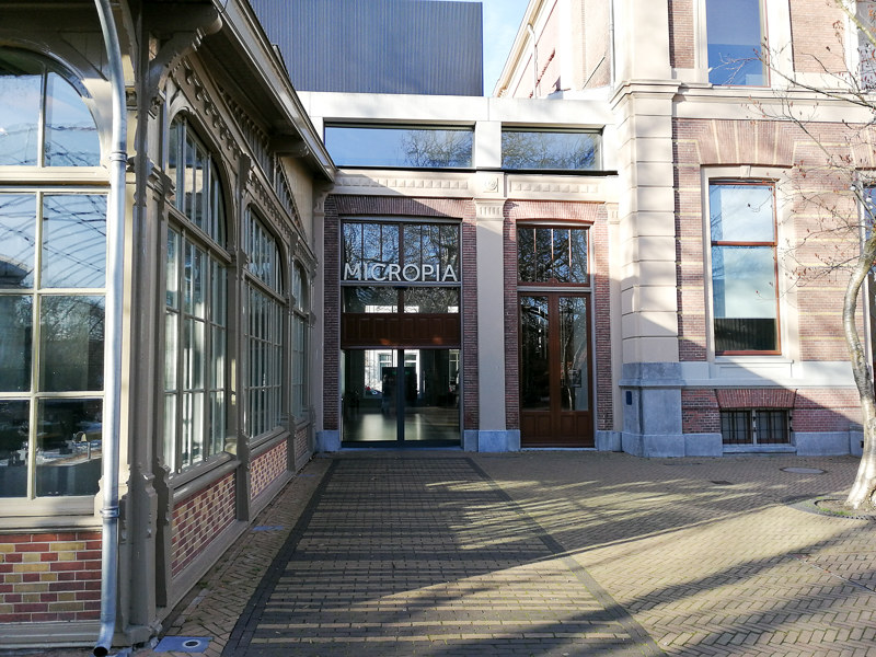 Buitenaanzicht van de ingang van het Micropia-gebouw met stenen gevel, boogramen en glazen deuren onder een bord met de tekst 'MICROPIA'