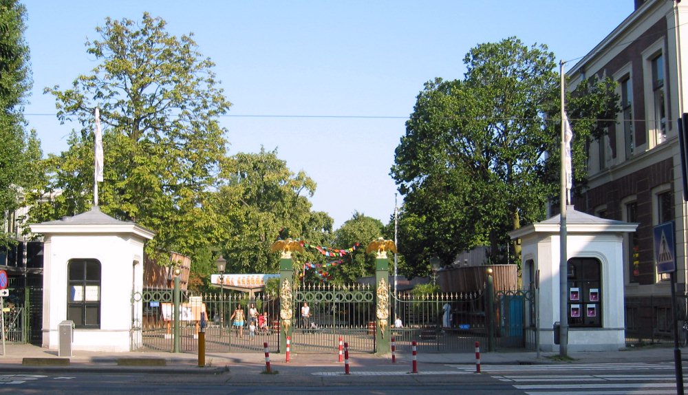 Gran puerta de entrada con forja ornamental flanqueada por casetas de portería blancas, árboles y visitantes en el zoo Artis