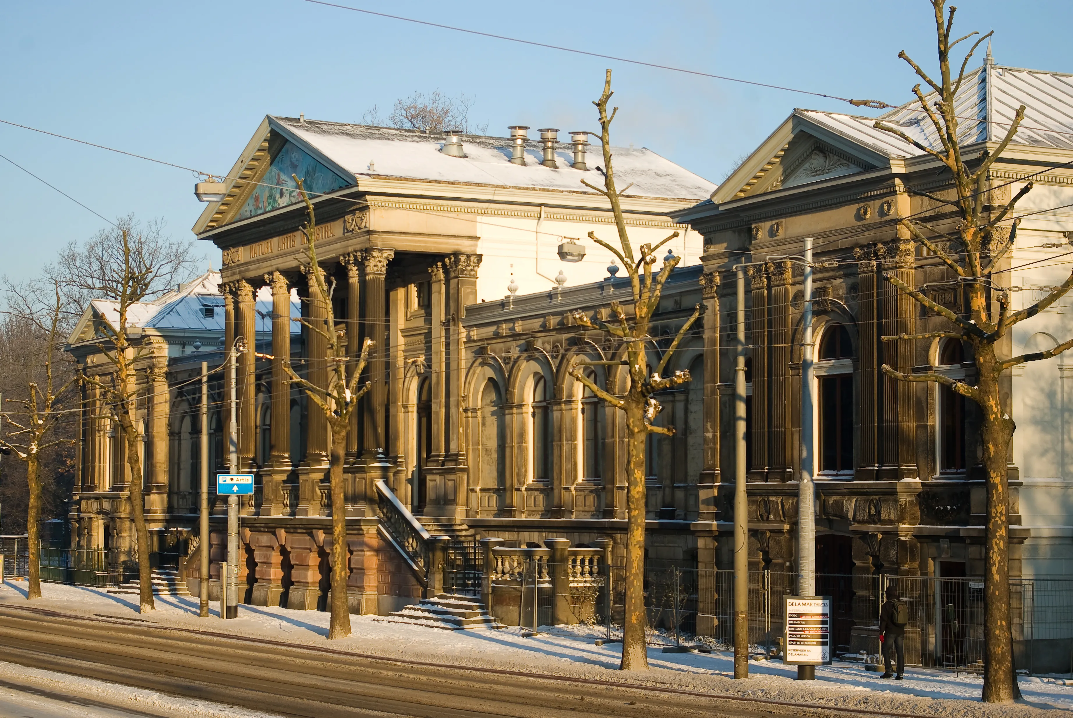 Un grandioso edificio histórico con arquitectura clásica, columnas y un tejado cubierto de nieve, rodeado de árboles desnudos y vías de tranvía.