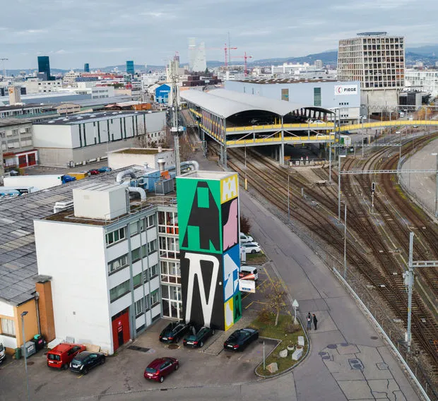 Vista aérea de un edificio industrial blanco que presenta un gran mural de grafiti colorido con letras estilizadas A y N en verde, negro, rosa y amarillo, situado junto a las vías del tren con un paisaje urbano de fondo