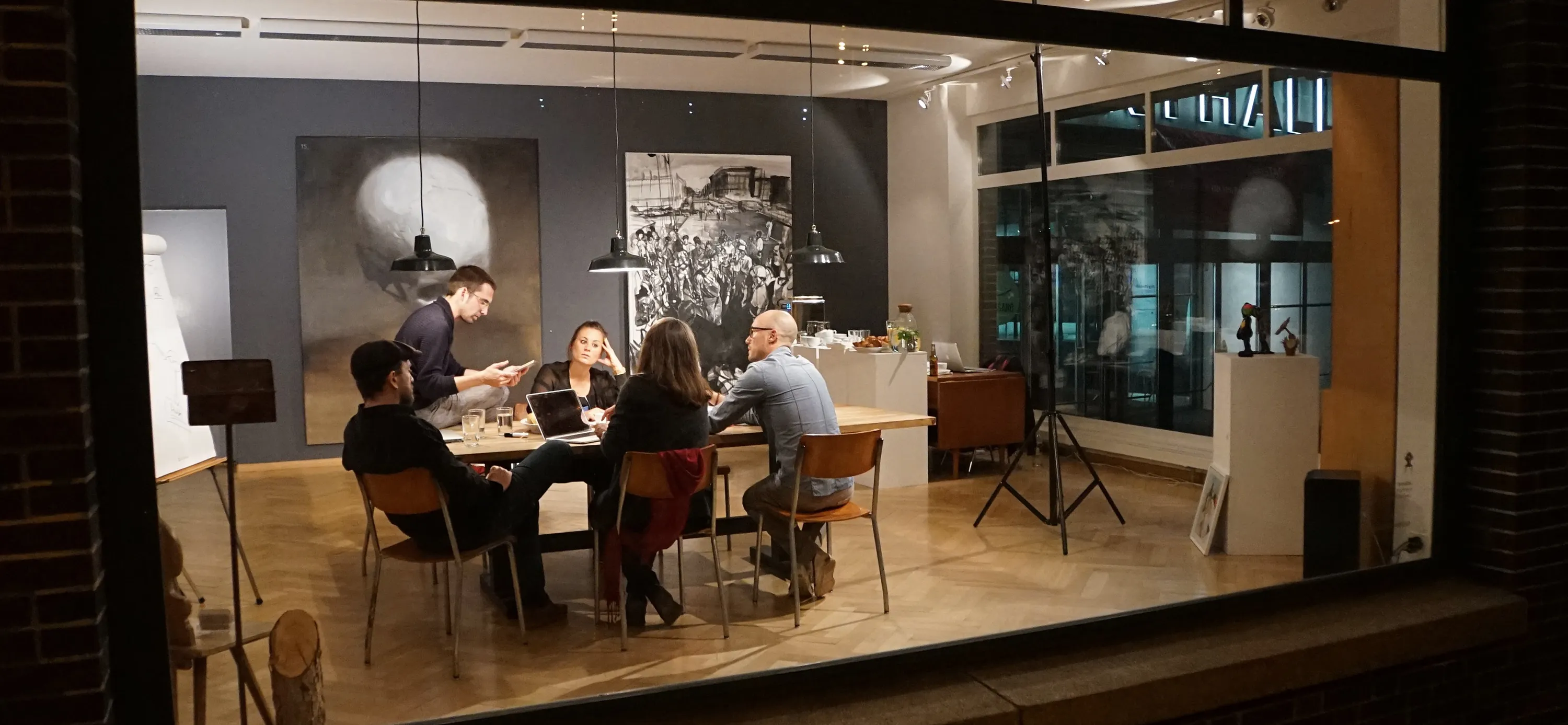 Personas sentadas alrededor de una mesa de madera en el interior de una galería por la noche, vistas a través de un gran escaparate, con una pintura circular grande y una fotografía en blanco y negro en las paredes