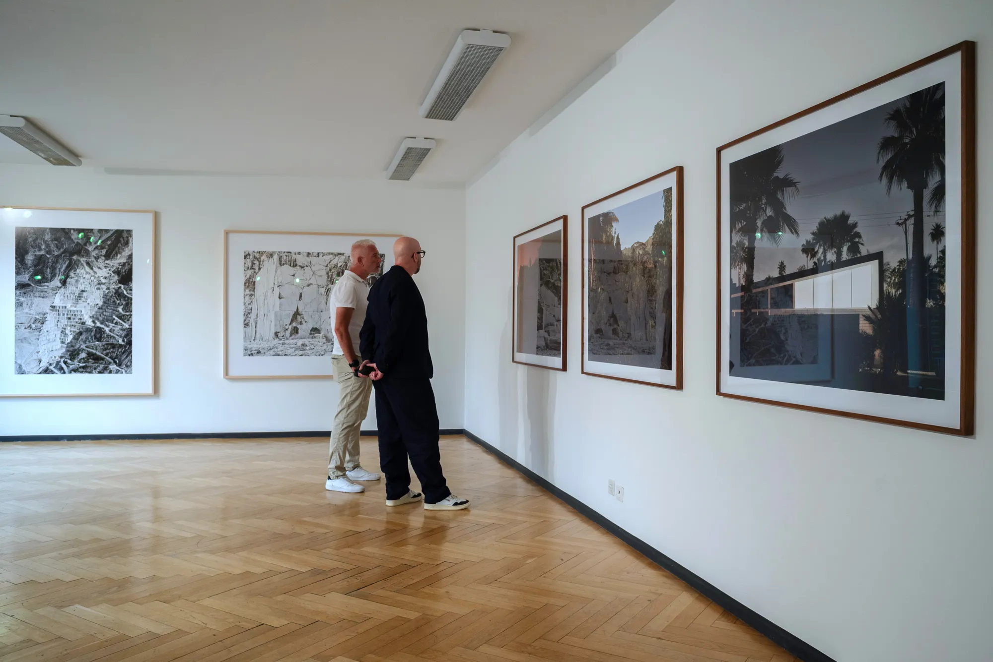 Dos hombres de pie en una galería observando grandes fotografías enmarcadas y obras de arte colgadas en paredes blancas sobre un suelo de parqué de madera.