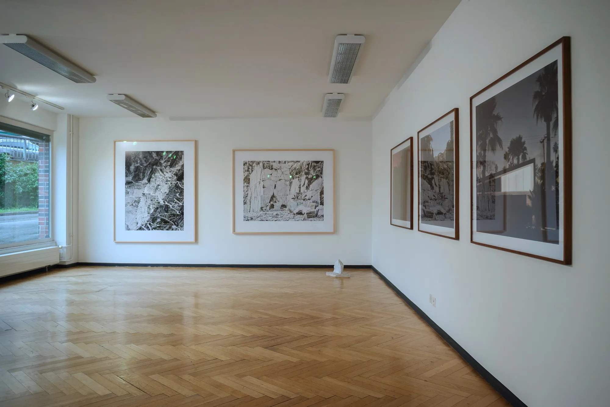 Interior de una galería con fotografías en blanco y negro enmarcadas sobre paredes blancas, suelo de parqué en espiga e iluminación de carril en el techo.