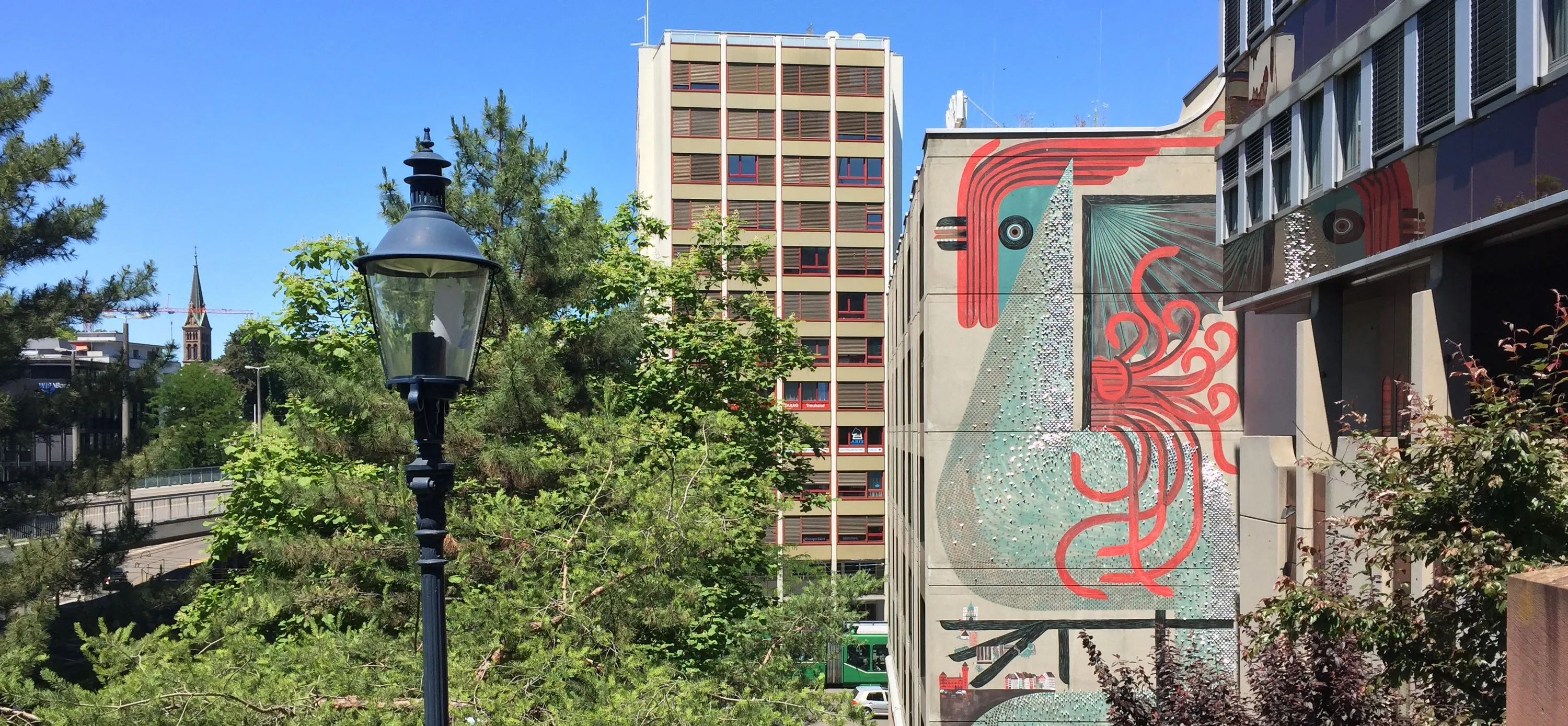 Un gran mural colorido de un pájaro estilizado en el costado de un edificio, con una farola negra y árboles verdes en primer plano bajo un cielo azul.