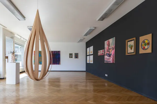Interior de una galería con una gran escultura colgante de madera en forma de lágrima, cuadros enmarcados en una pared azul marino y otra blanca, con suelo de parqué de madera e iluminación fluorescente en el techo.