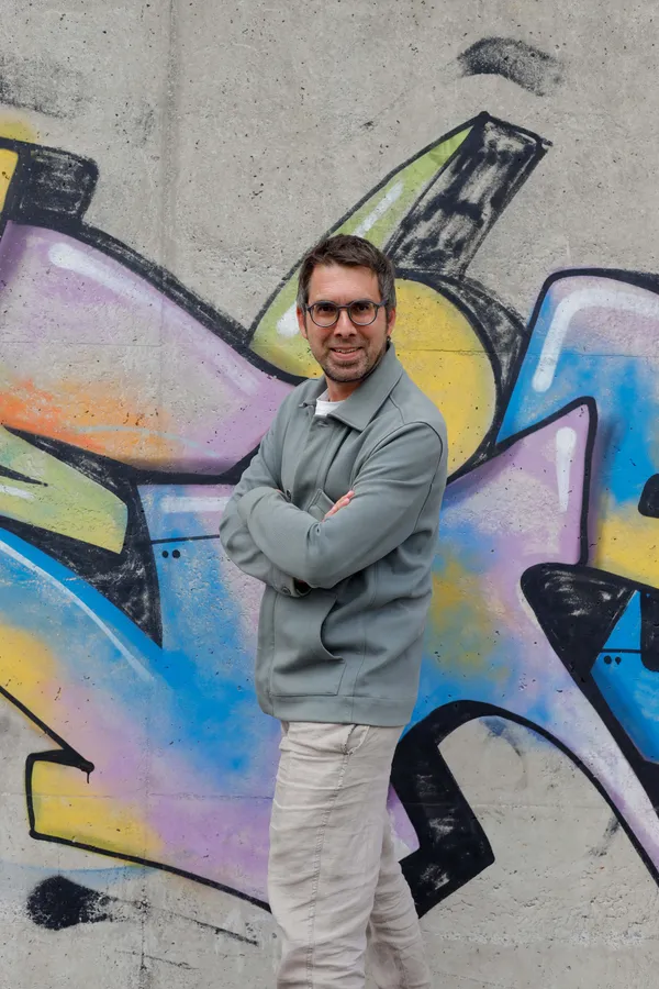 Un hombre sonriente con gafas y chaqueta gris posa con los brazos cruzados frente a una pared de hormigón cubierta de coloridos grafitis.