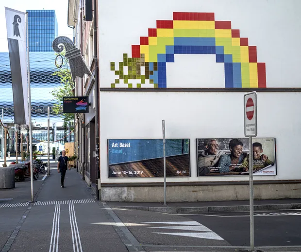 Vista de la calle de una fachada blanca con un gran mural de arte pixelado de un arcoíris y un personaje de space invader verde, pósteres de Art Basel en la parte inferior de la pared y un peatón en la acera
