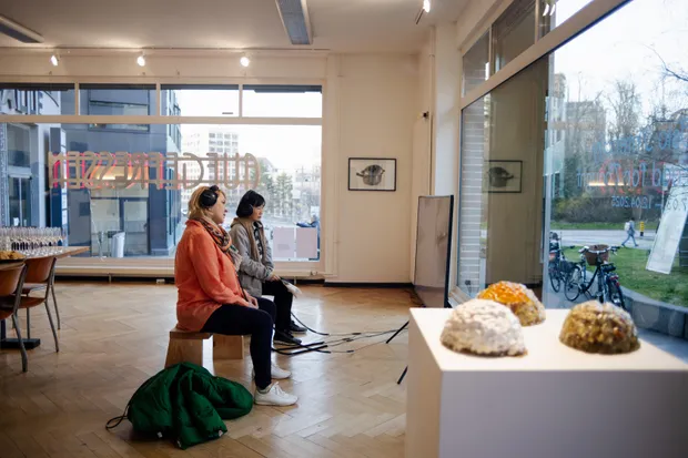 Interior de la galería Artstübli con dos visitantes sentados en taburetes cerca de grandes ventanales con textos gráficos, objetos escultóricos sobre un pedestal blanco en primer plano y obras de arte enmarcadas en las paredes.