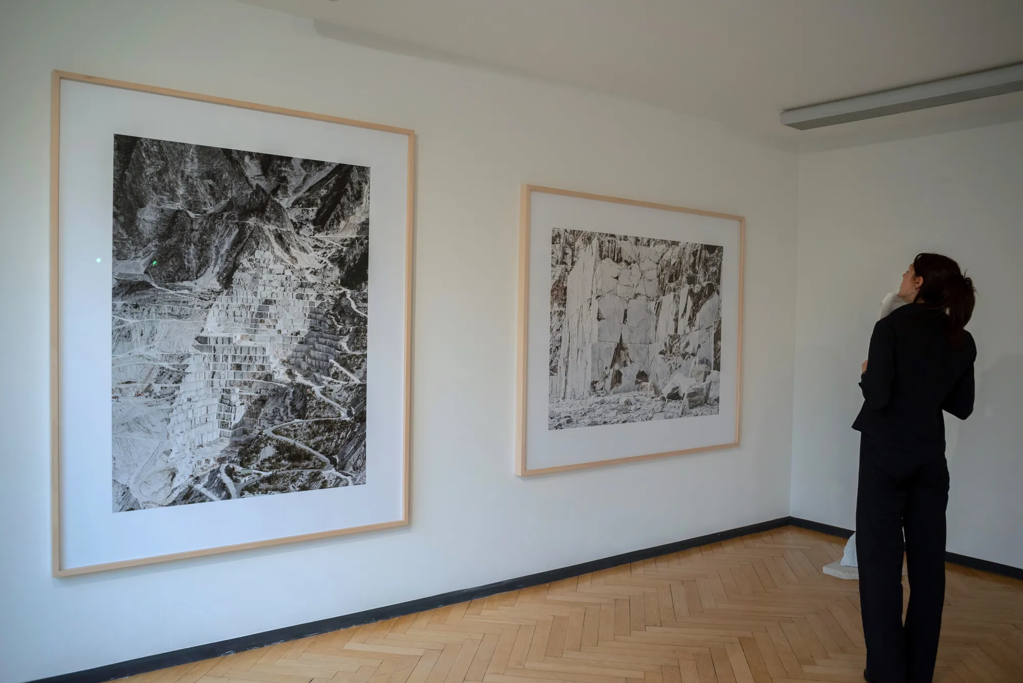 Dos grandes obras de arte en blanco y negro enmarcadas cuelgan de una pared blanca de una galería mientras una mujer las observa.