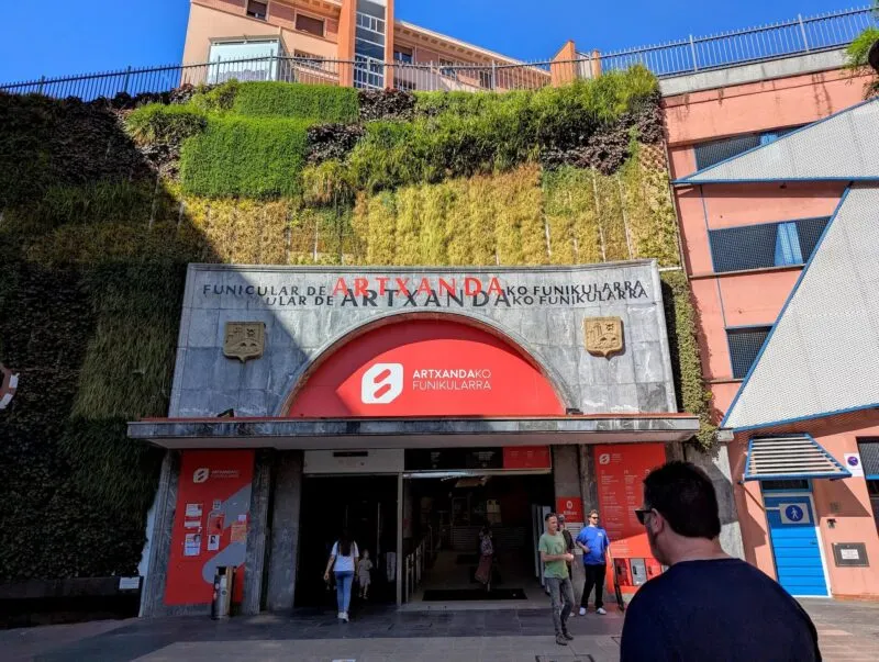 La fachada de entrada del Funicular de Artxanda en Bilbao, con una entrada de arco rojo que lleva el logotipo de Artxanda, una gran pared vegetal arriba, letreros de piedra que dicen 'Funicular de Artxanda' y gente entrando y saliendo.