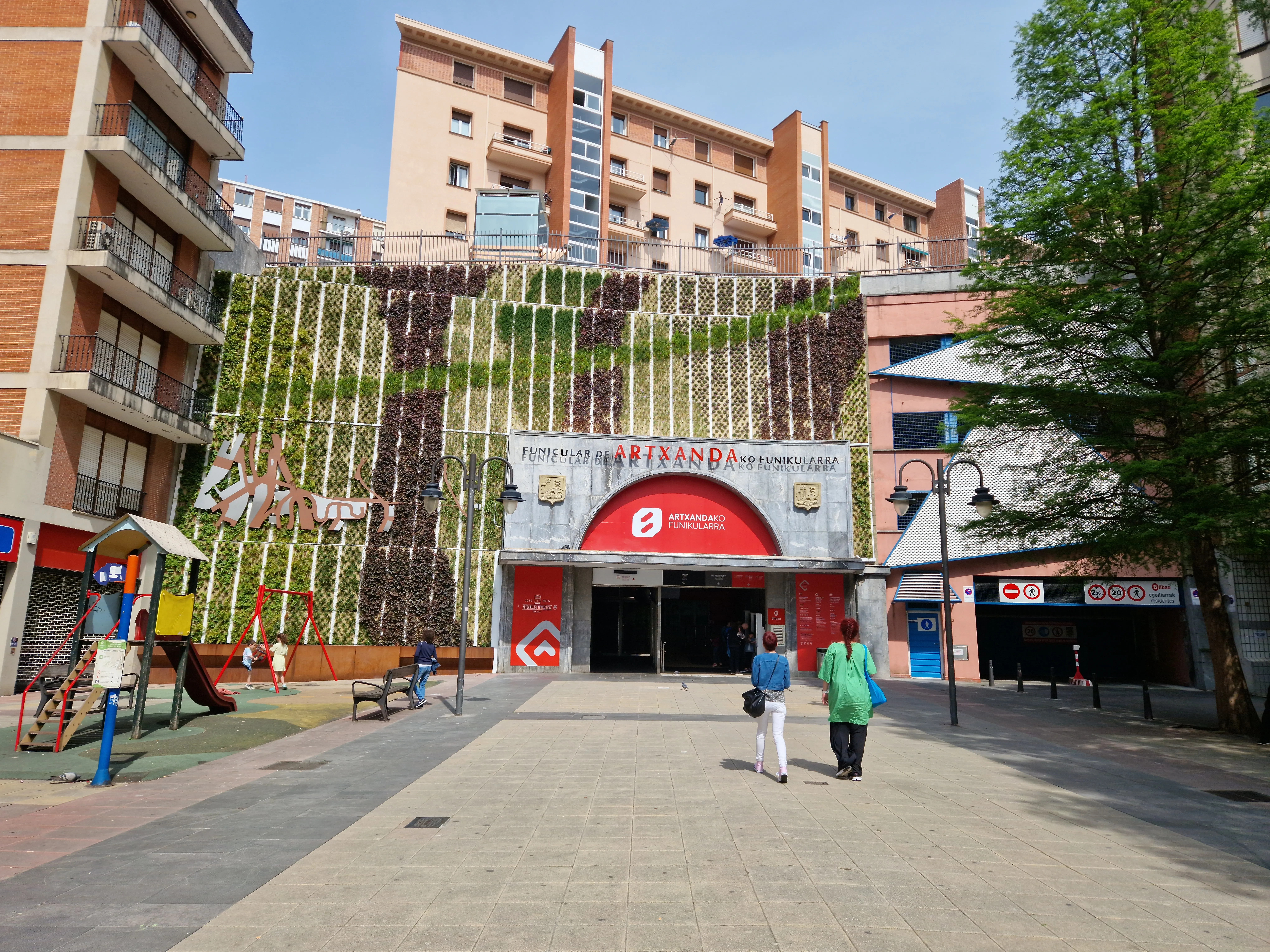Vista exterior de la entrada de la estación del Funicular de Artxanda con el arco rojo, un mural de jardín vertical y los edificios residenciales circundantes en Bilbao