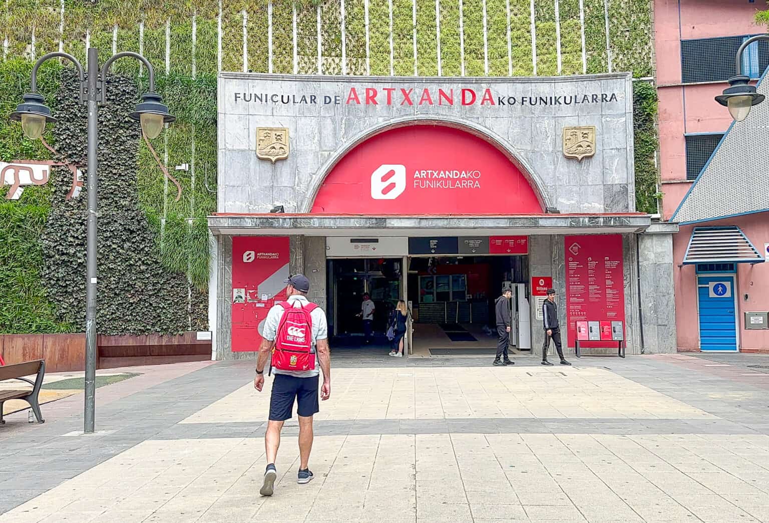 Vista exterior de la entrada del Funicular de Artxanda con un gran cartel arqueado de color rojo en el que se lee 'Funicular de Artxanda' y una persona con una mochila roja caminando hacia la entrada