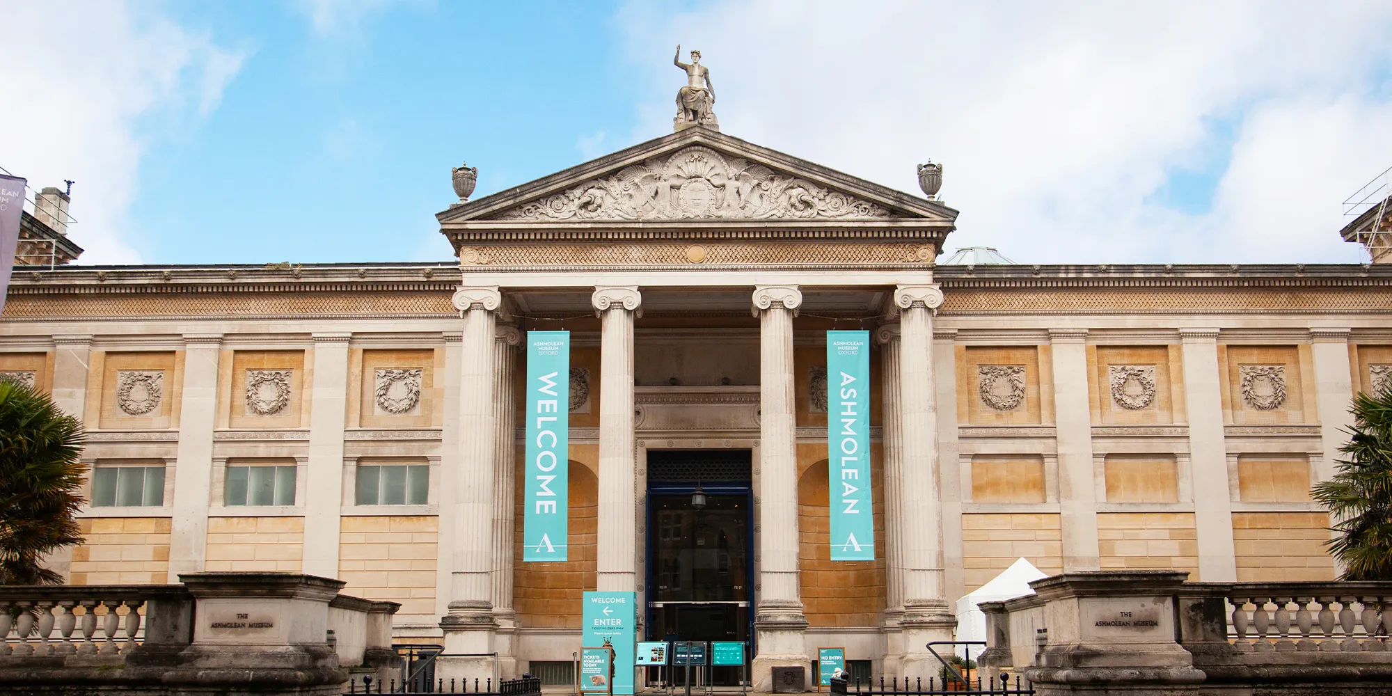 Neoklasycystyczna fasada Ashmolean Museum z kolumnowym portykiem, trójkątnym frontonem ze zwieńczającą go rzeźbą oraz dwoma turkusowymi banerami z napisami MUSEUM i ASHMOLEAN zawieszonymi między kolumnami.
