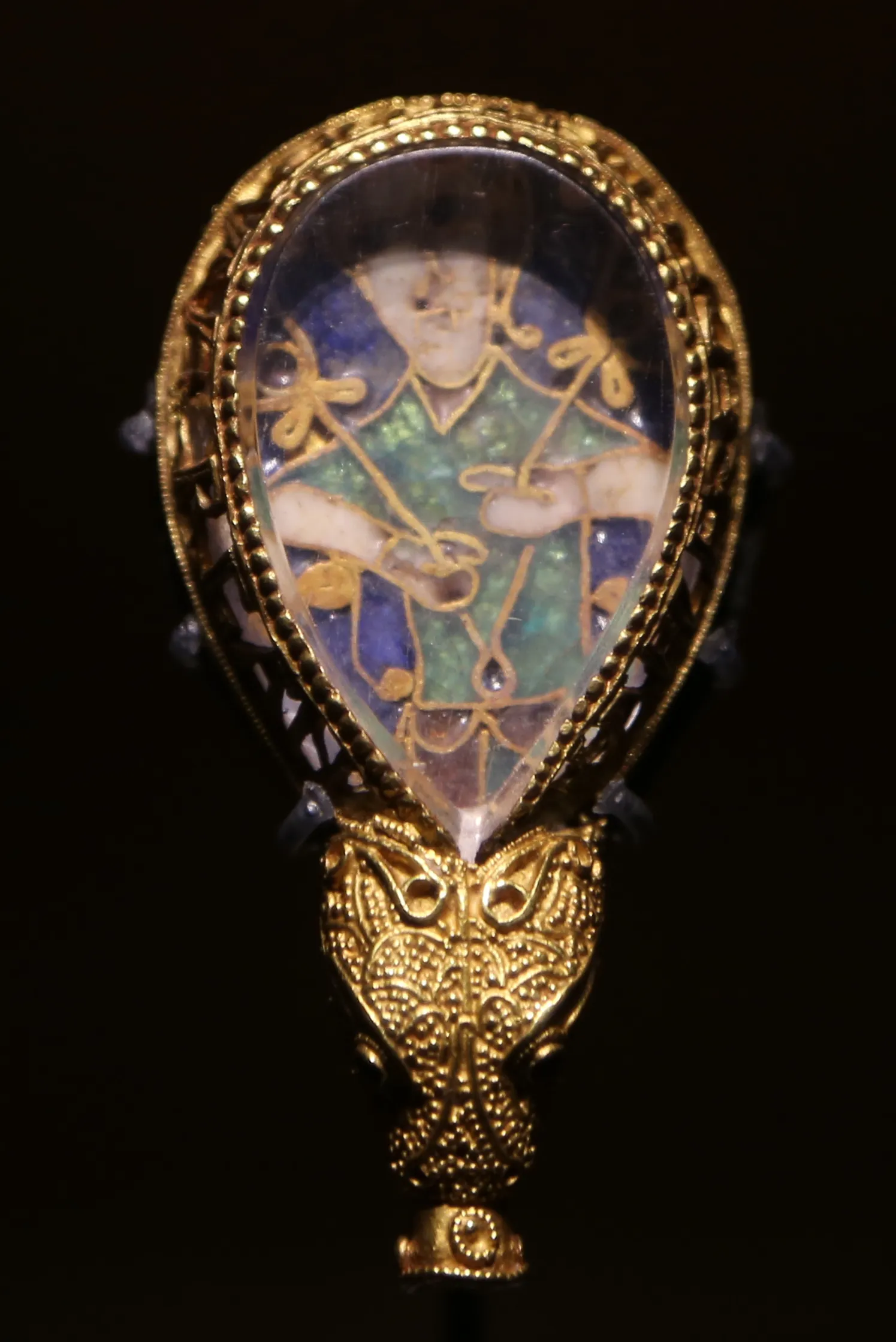 The Alfred Jewel, kunsztowny anglosaski artefakt ze złota i emalii z nakrywką z kryształu górskiego, przedstawiający postać w szatach z zielonej i niebieskiej emalii, oprawiony w złotą filigranową ramę w kształcie łzy z zakończeniem w formie zwierzęcej głowy, na czarnym tle
